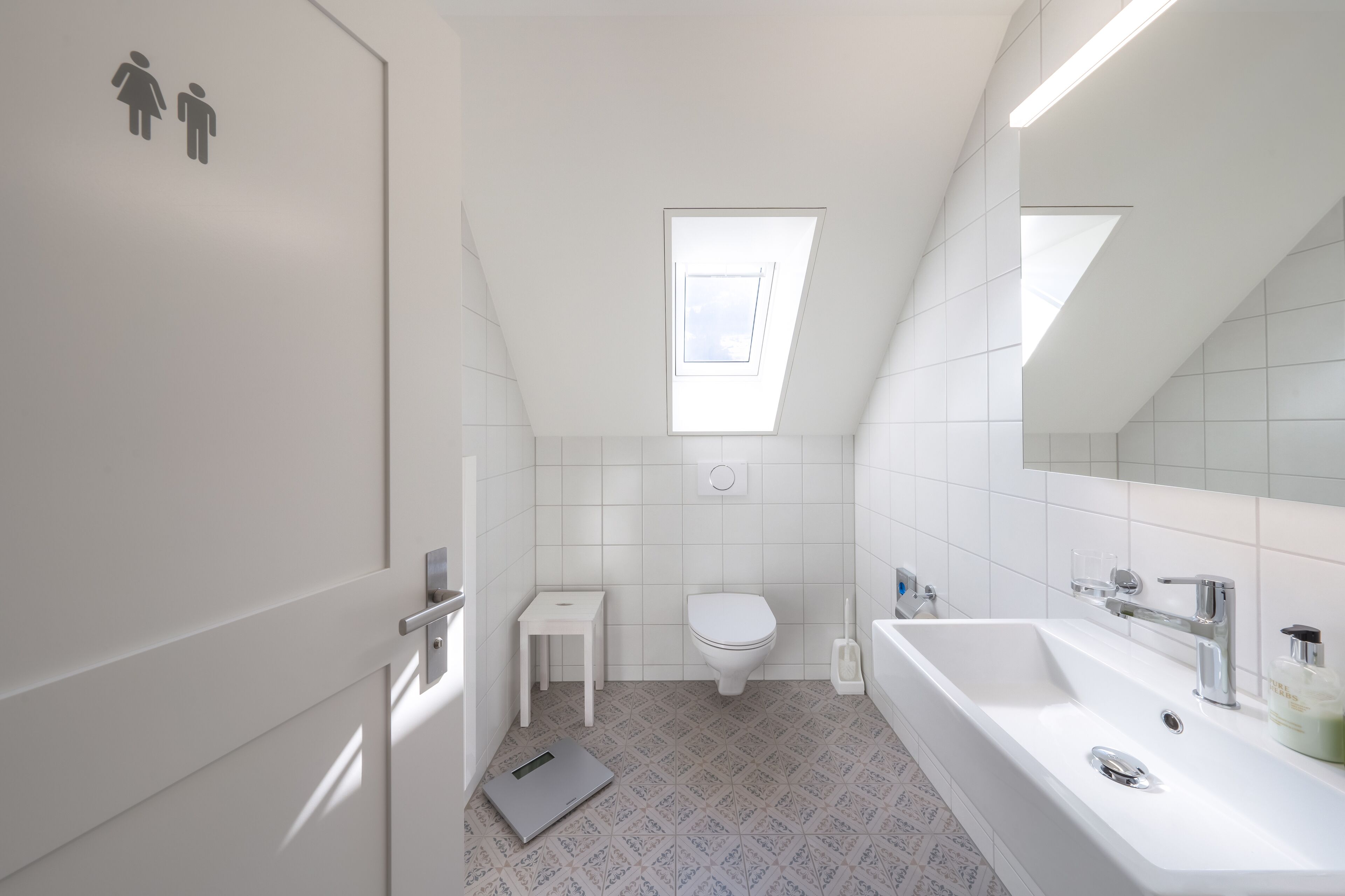 Une salle de bain moderne avec un puits de lumière, des carreaux blancs, des toilettes, une petite table et une baignoire.
