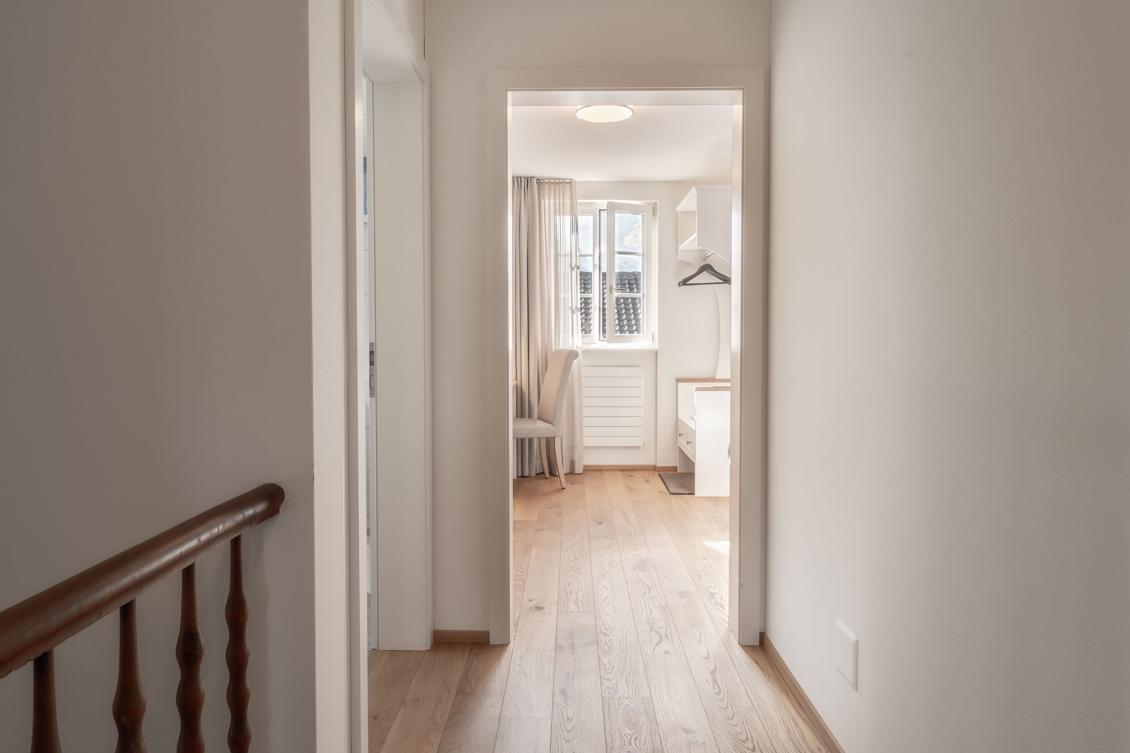 Un pasillo con barandilla de madera que conduce a una habitación con ventana, una silla y un perchero.