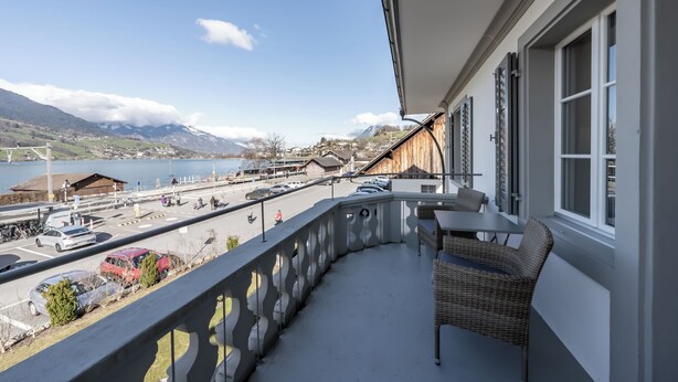 A balcony with a view of a lake and mountains, a table with chairs, and a parking area with cars.