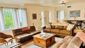 A cozy living room with a brown leather sofa, a wooden coffee table with books, and a large window with curtains. - The Riverpath Inn, just steps from the Willamette river & Rose Gardens, Eugene (Eugene)