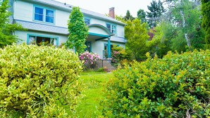 A two-story house with a front porch, surrounded by lush greenery and flowering plants. - The Riverpath Inn, just steps from the Willamette river & Rose Gardens, Eugene (Eugene)