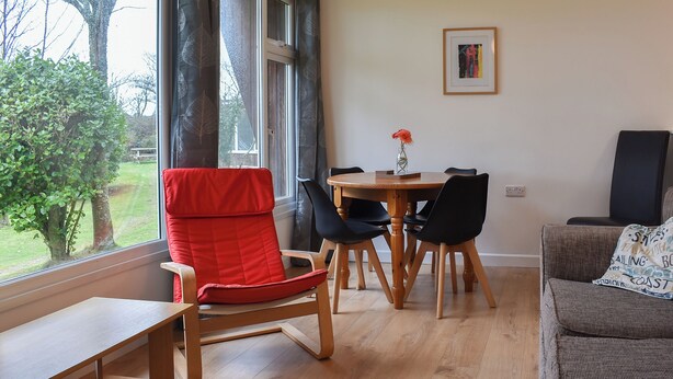 A dining area with a wooden table, black chairs, and a red chair. There is a window with curtains, a framed picture on the wall, and a sofa with patterned cushions. - 2 bedroom accommodation in Mount Hawke, near Redruth (Mount Hawke, near Redruth)