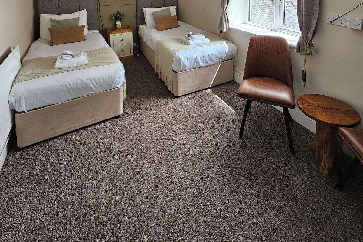 A hotel room with two single beds, a small wooden table, a chair, a window with curtains, and a ceiling light.