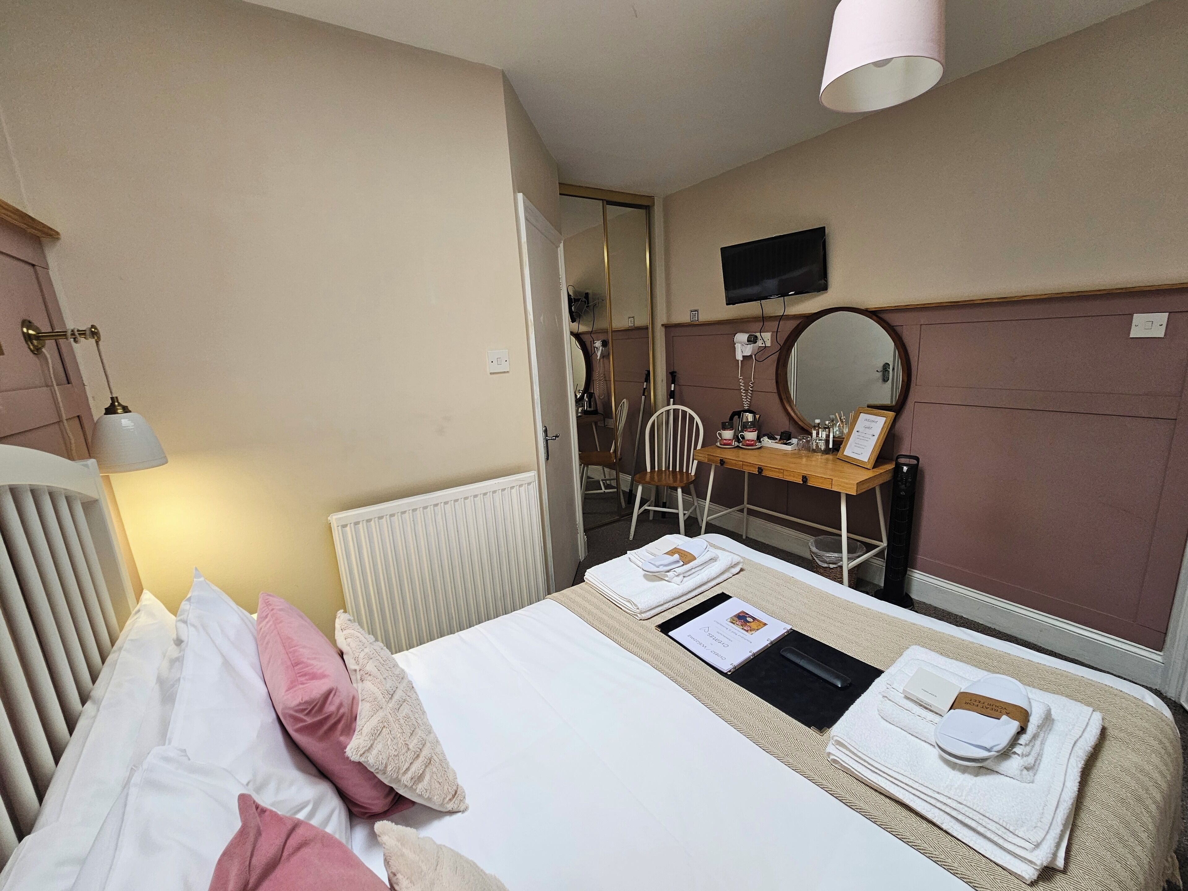 A hotel room with a bed, a desk with a mirror, a chair, and a radiator.