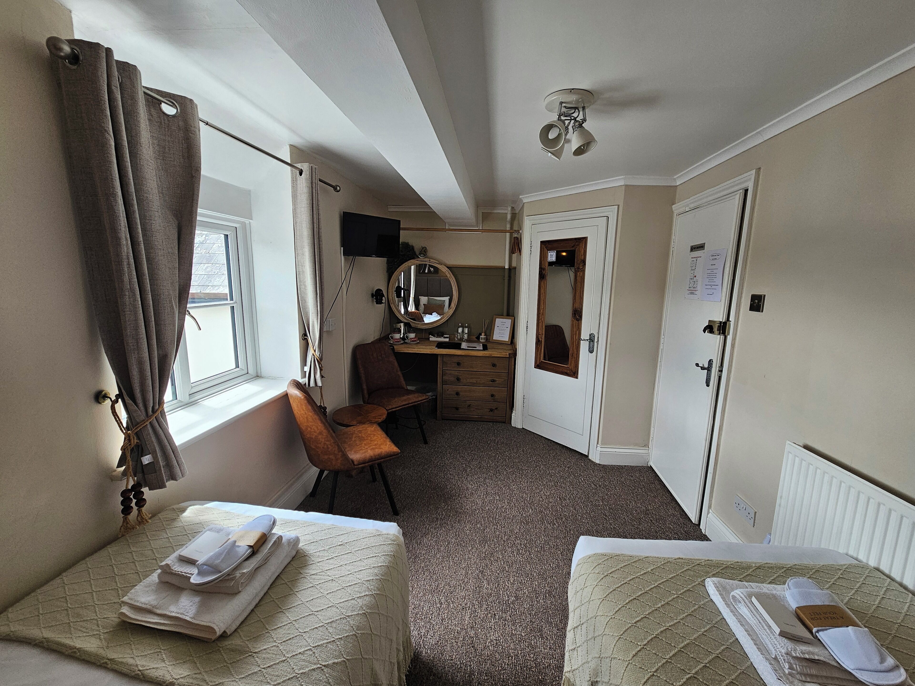 A hotel room with two single beds, a desk with a chair, a mirror, and a wardrobe.