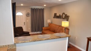 A living room with a brown leather sofa, a kitchen area with granite countertops, and a wall-mounted lamp. - 2BA 6-Beds 10-min to downtown Nashville! (Nashville)