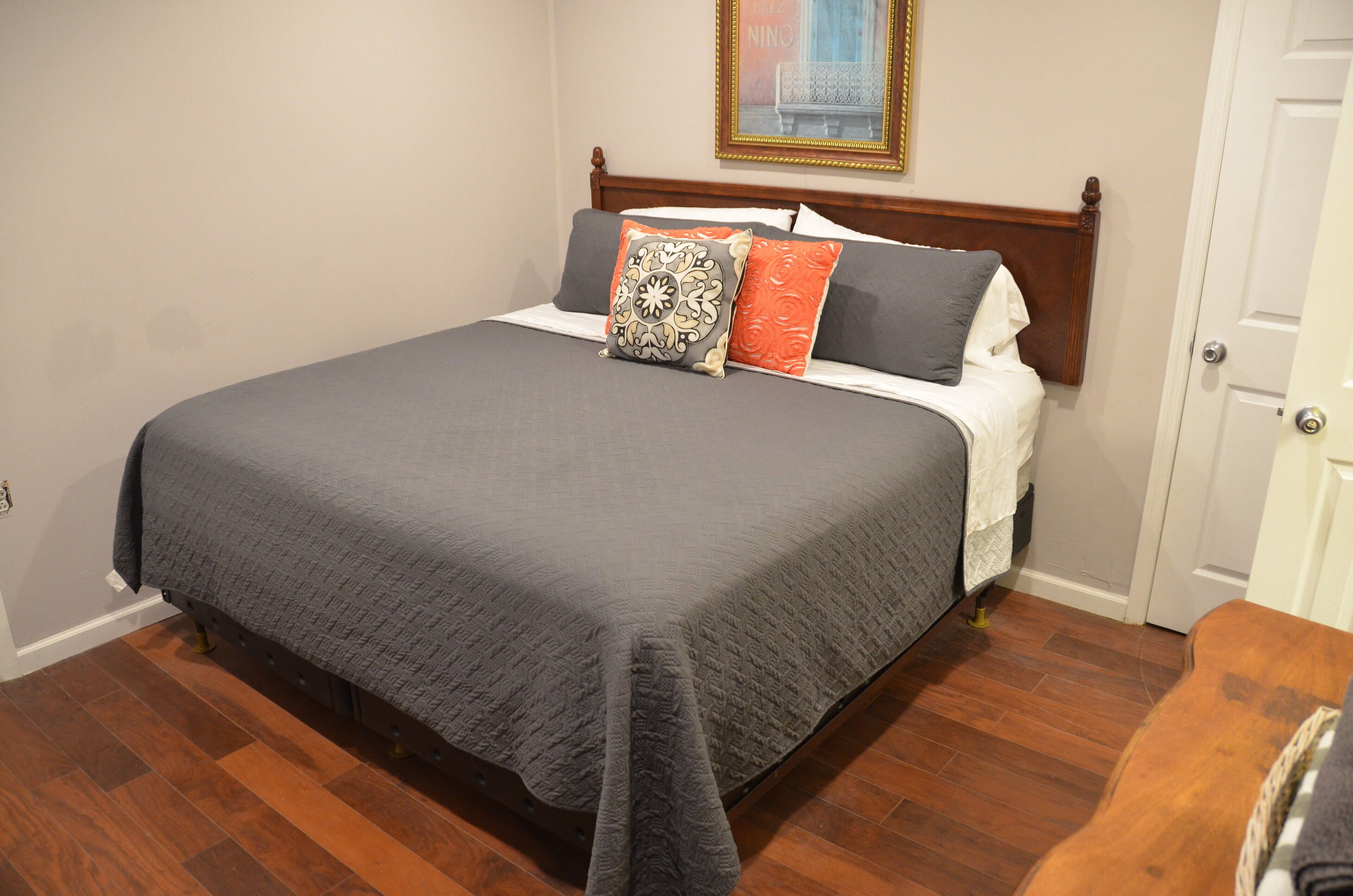 Una cama bien hecha con colcha gris, dos almohadas con diseño estampado y cabecera de madera.