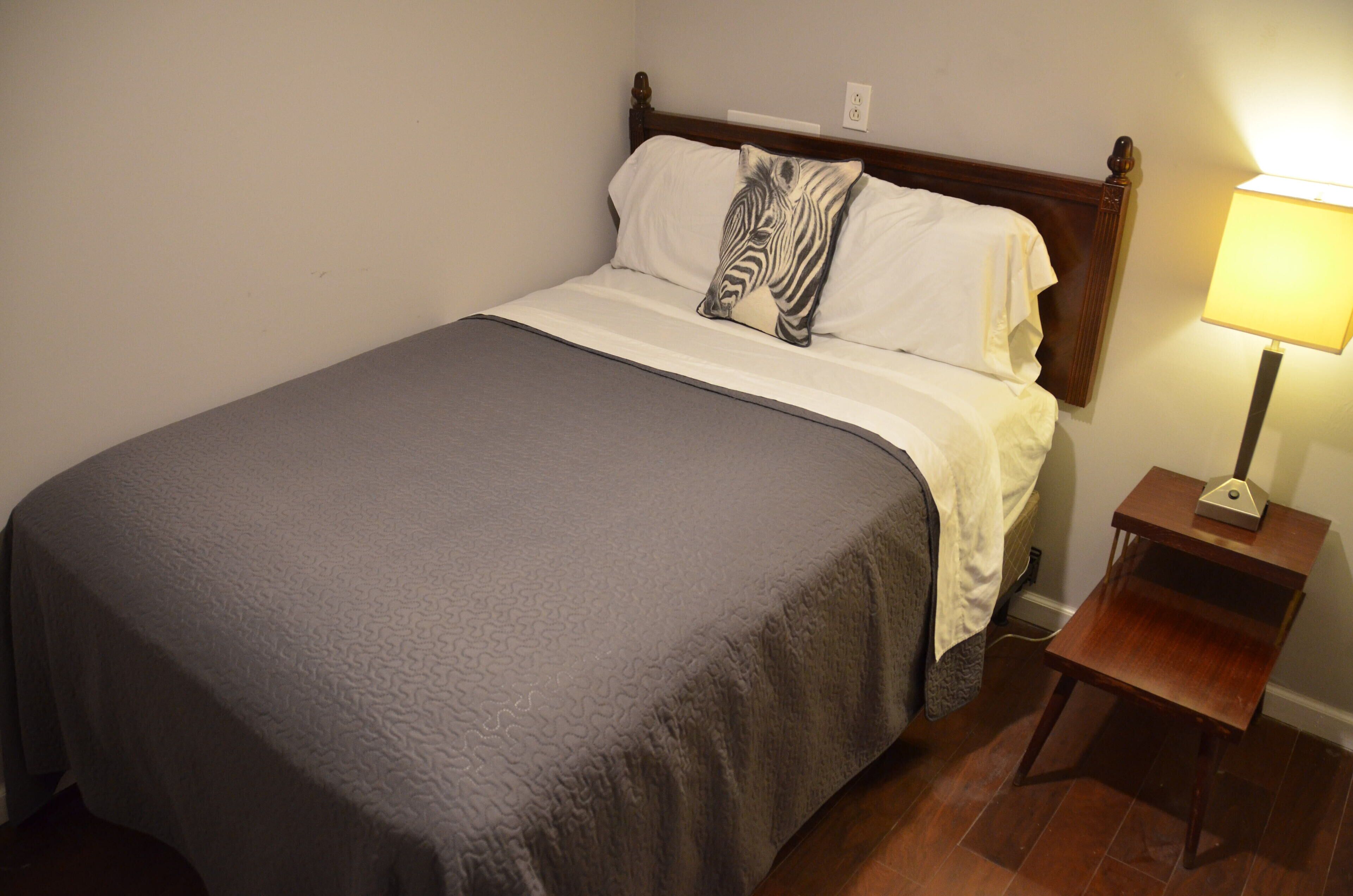 Una cama bien hecha con un cojín estampado de cebra, una mesita de noche con lámpara y un cabecero de madera.