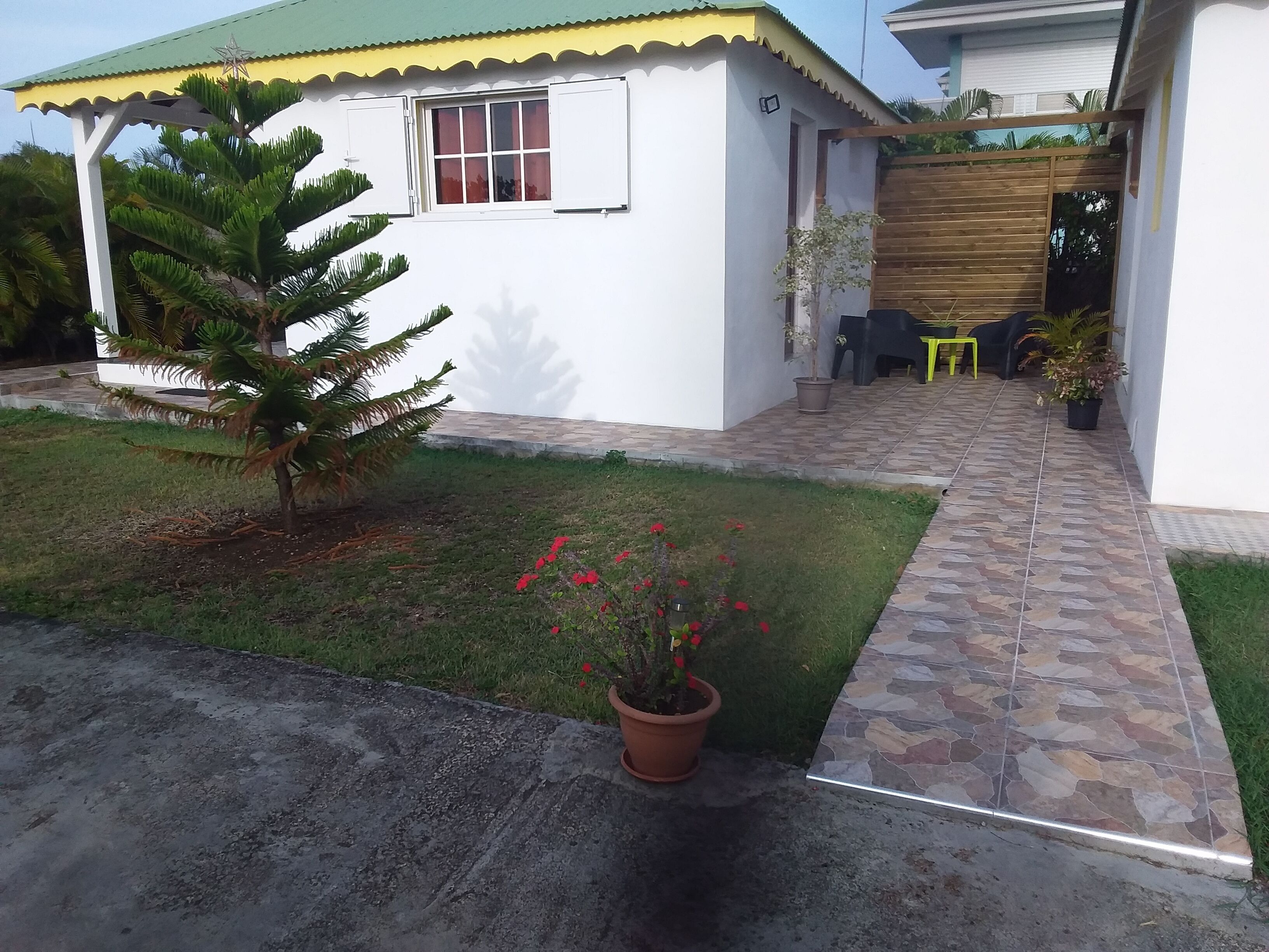Een klein huis met een betegeld terras, een kleine tuin met een palmboom en potplanten, en een houten omheining.