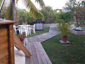 Una terraza de madera que conduce a una mesa y sillas blancas de plástico al aire libre, con una parrilla y plantas en macetas cerca.