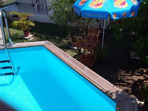 A swimming pool with a wooden deck, surrounded by a garden with potted plants and a table with chairs. - Comfortable f2 gite with swimming pool (Le moule)