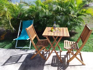 A wooden table and four chairs set outdoors with a pool and palm trees in the background. - Comfortable f2 gite with swimming pool (Le moule)