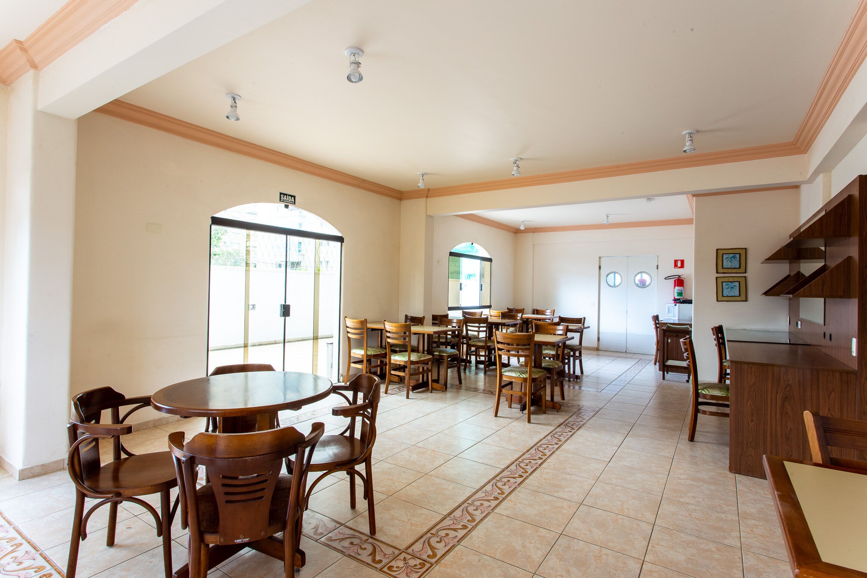 A spacious dining area with multiple wooden tables and chairs, a kitchen area with wooden cabinets, and a view of the outside through large windows.