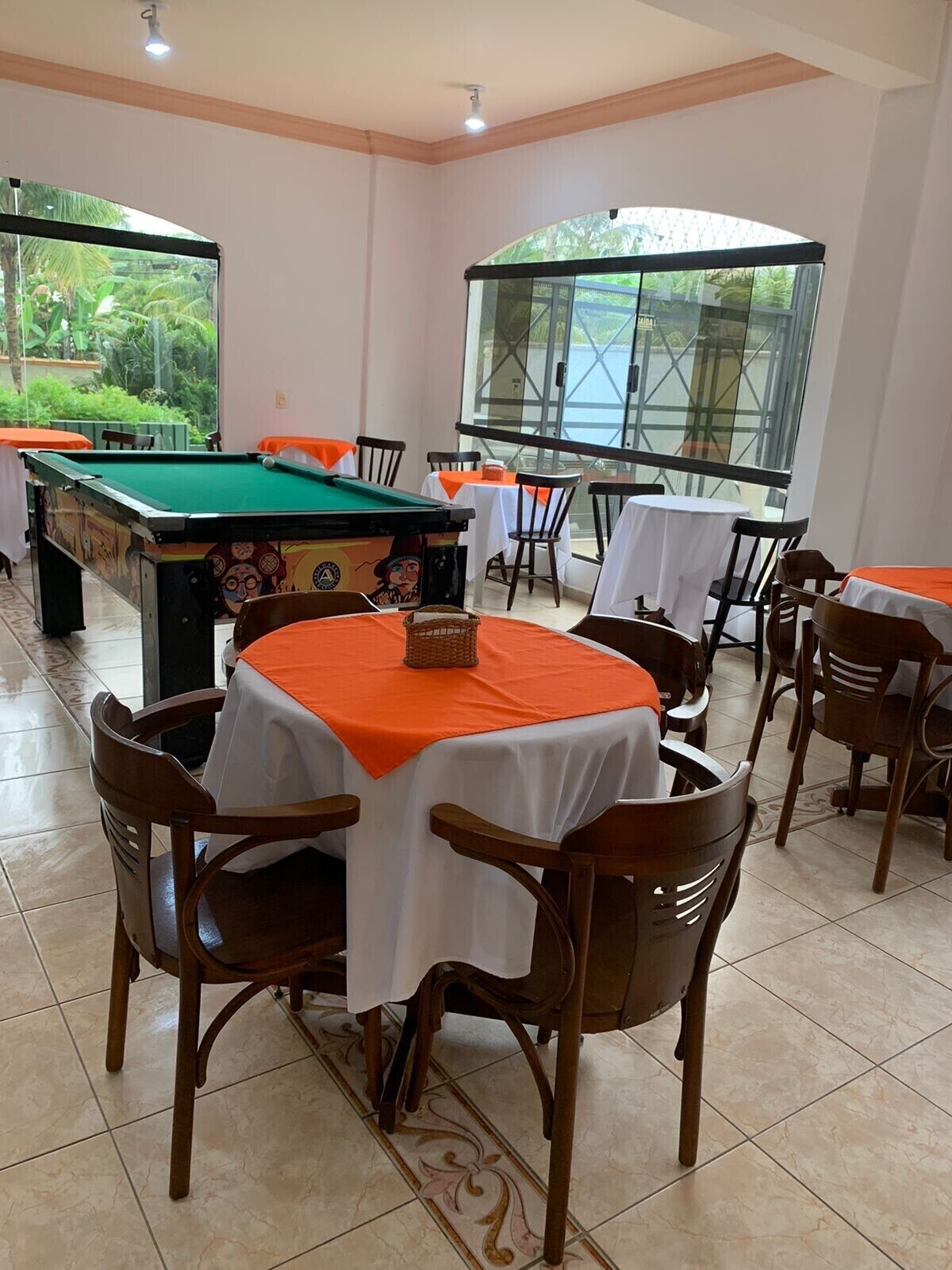 Een poolzaal met meerdere tafels en stoelen, een pooltafel en grote ramen met uitzicht op groen.