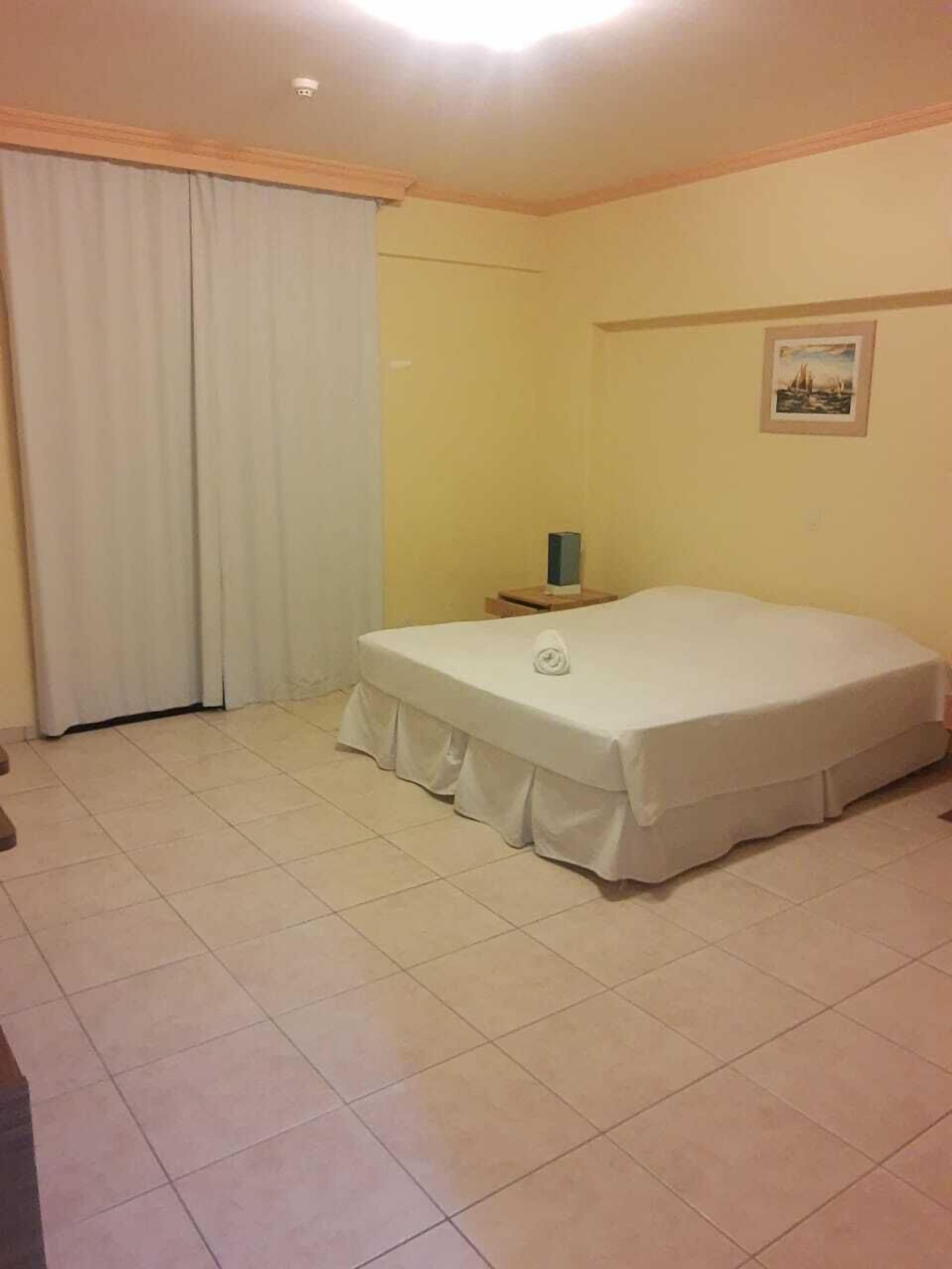 Una cama individual con colcha blanca, una mesita de noche con lámpara y una ventana con cortinas.