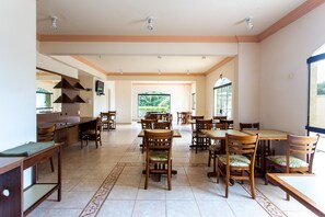 Un comedor amplio con múltiples mesas y sillas de madera, una zona de cocina con mostrador y electrodomésticos, y ventanas grandes que dejan entrar luz natural.