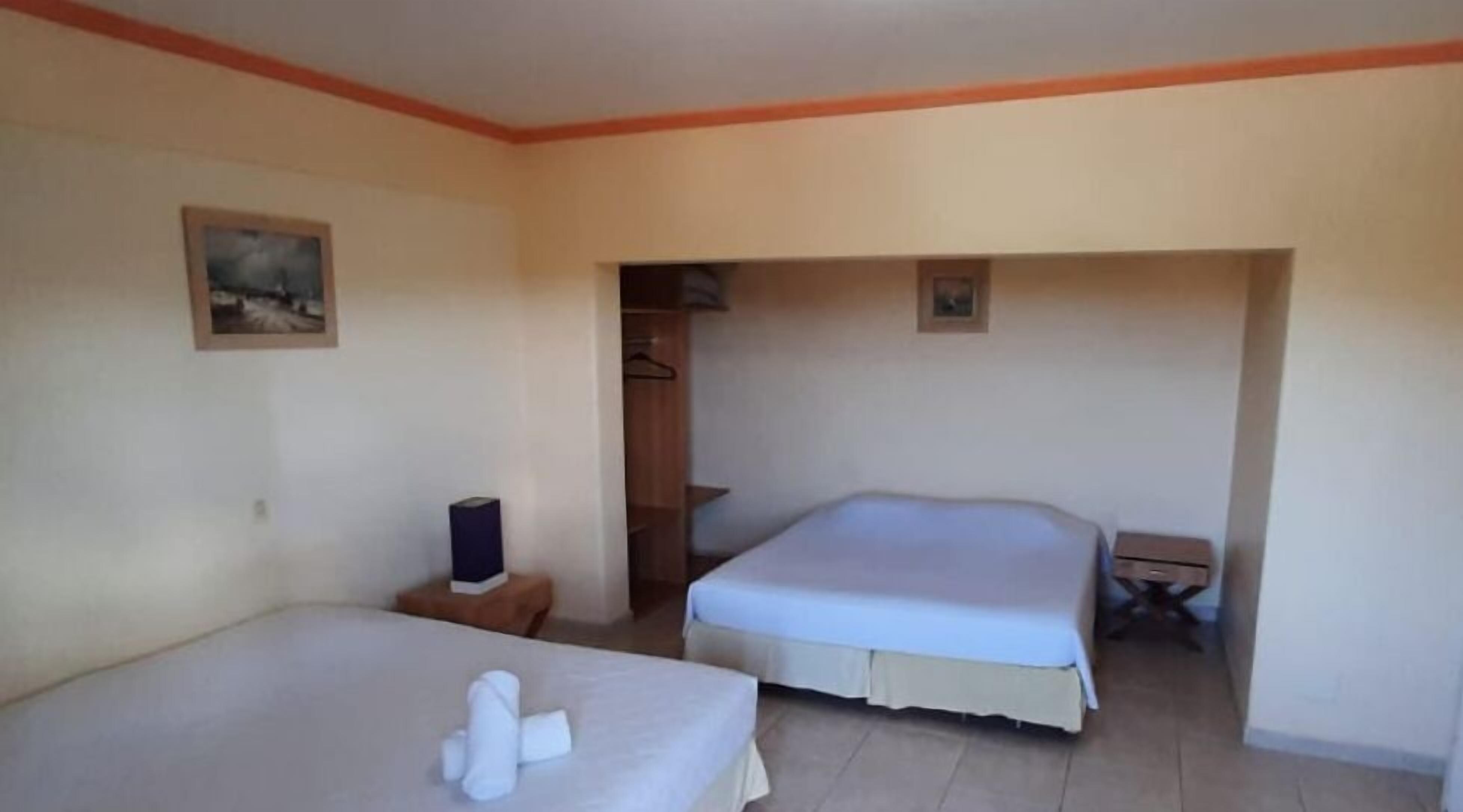 A hotel room with two beds, a small table, a wardrobe, and a painting on the wall.