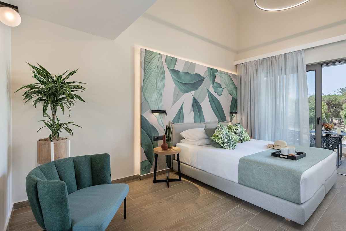 A modern hotel room with a bed, a green armchair, a potted plant, and a large window with curtains.