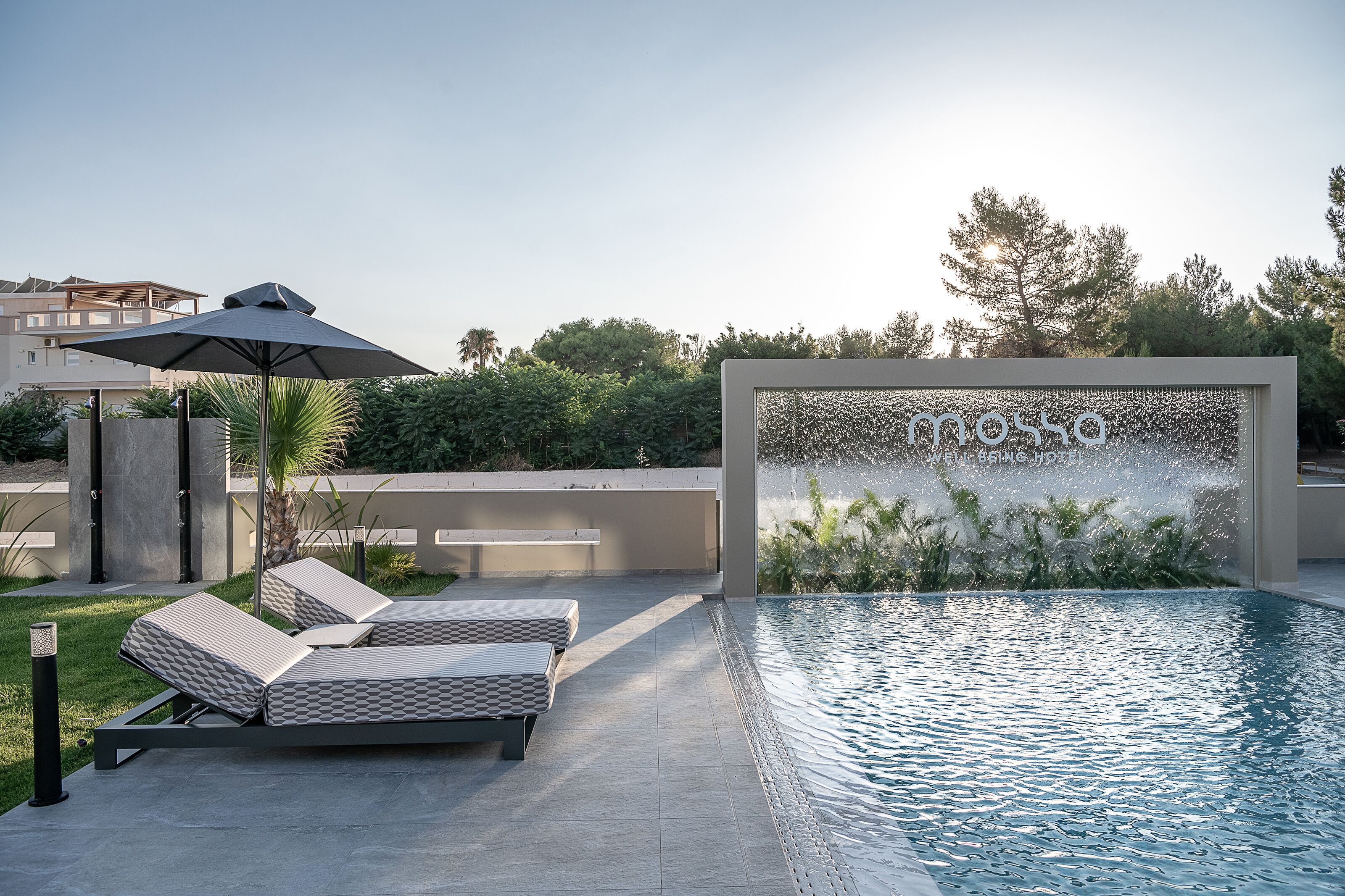 Un área de piscina exterior moderna con sillones de descanso, una fuente de agua y cielo despejado.