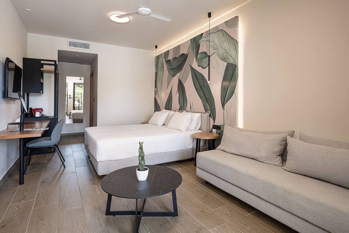 A modern hotel room with a bed, a sofa, a small table with a plant, a desk, and a TV.
