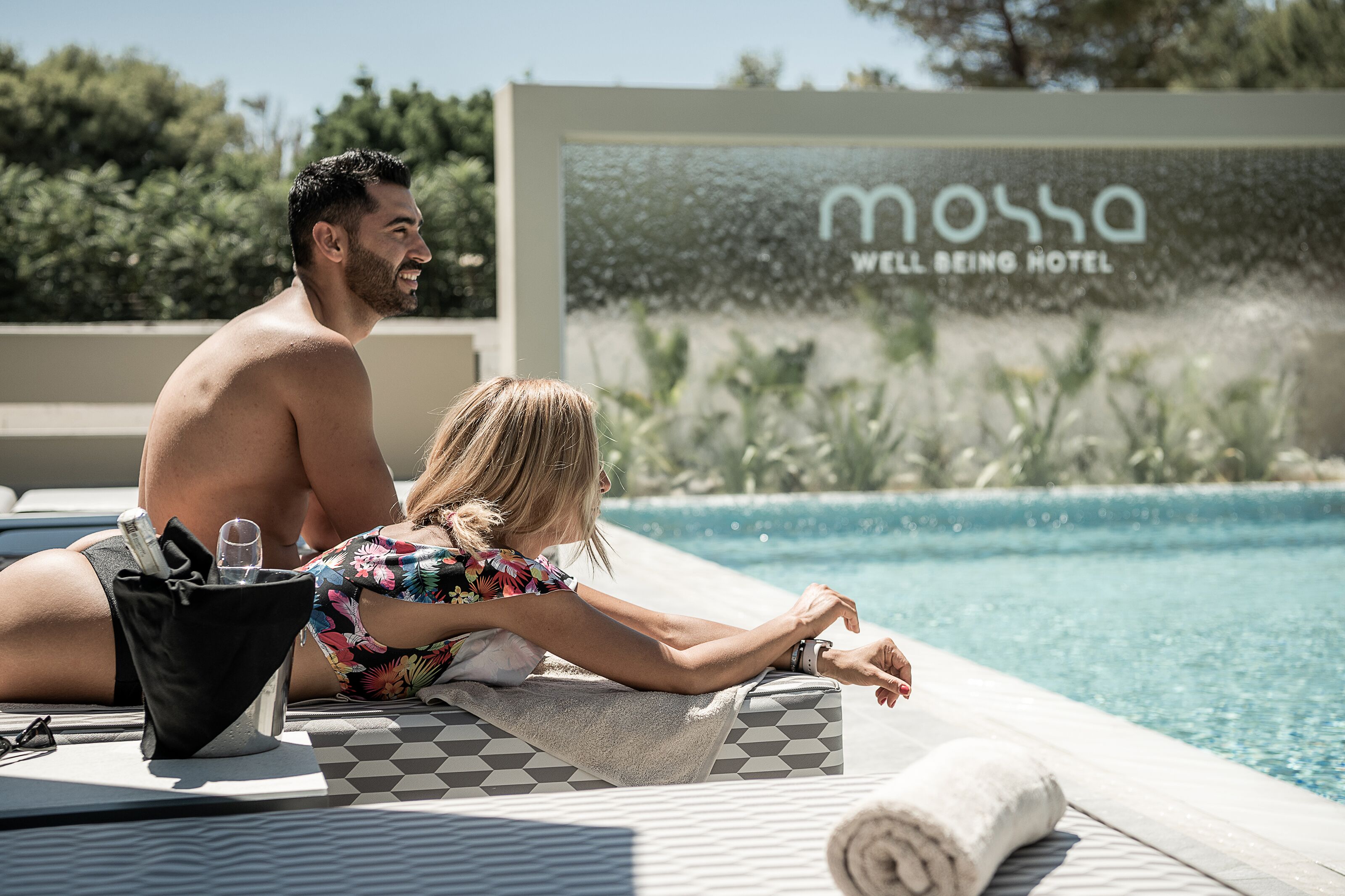 Una pareja se relaja junto a una piscina, con un cartel que dice "Mossa Well Being Hotel" al fondo.