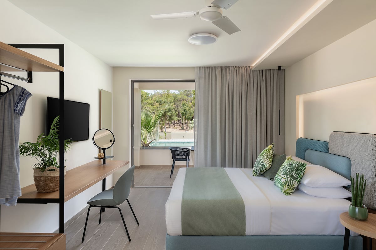 A modern hotel room with a bed, bedside table, chair, TV, and a view of the outdoors.