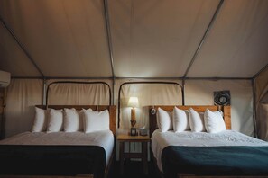 A tented room with two beds, a nightstand with a lamp, and a small table.