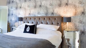 Ein Schlafzimmer mit einer gesteppten Kopfteil, einem Bett mit weißen Leinen, einem schwarzen Zierkissen und einer Wand mit Waldfoto.