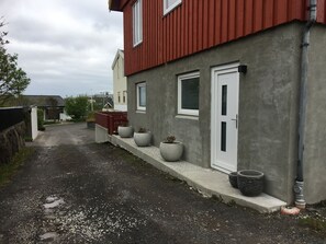 A narrow driveway leading to a house with a red roof and grey walls, potted plants along the side, and a concrete pathway.