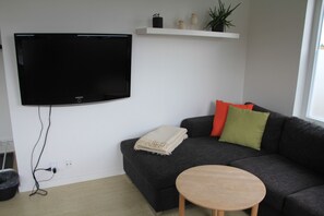 A modern living room with a flat-screen TV mounted on the wall, a grey sofa with colorful cushions, a small wooden table, and a white shelf above the sofa.