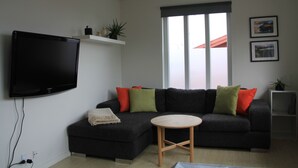 A modern living room with a sectional sofa, a flat-screen TV, a small round table, and a window with a view of a red-roofed building. - Nordic Inn Thorshavn (Tórshavn)