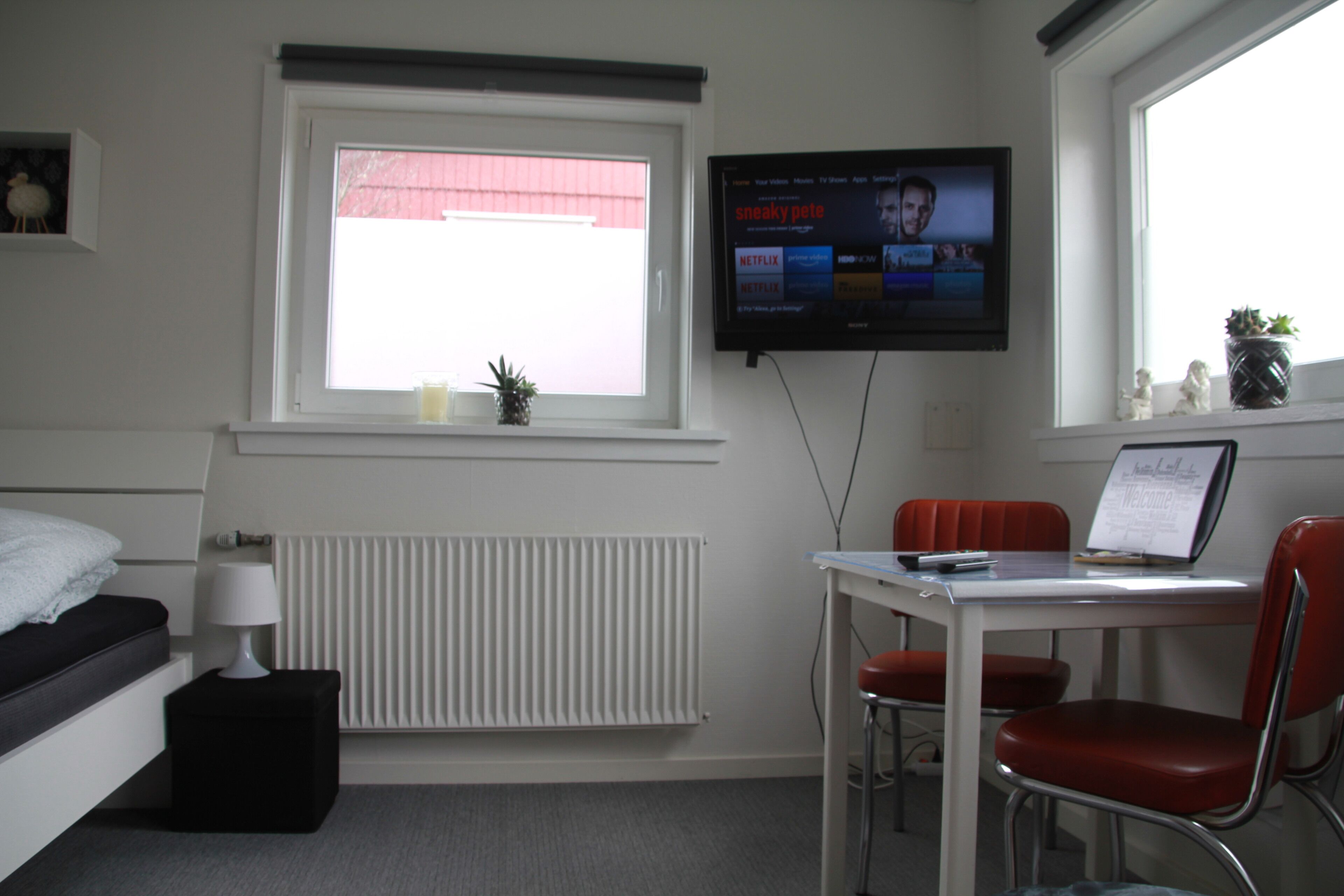 A hotel room with a bed, bedside table, lamp, window with blinds, radiator, desk with two chairs, and a TV mounted on the wall.