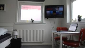 A hotel room with a bed, bedside table, lamp, window with blinds, radiator, desk with two chairs, and a TV mounted on the wall.