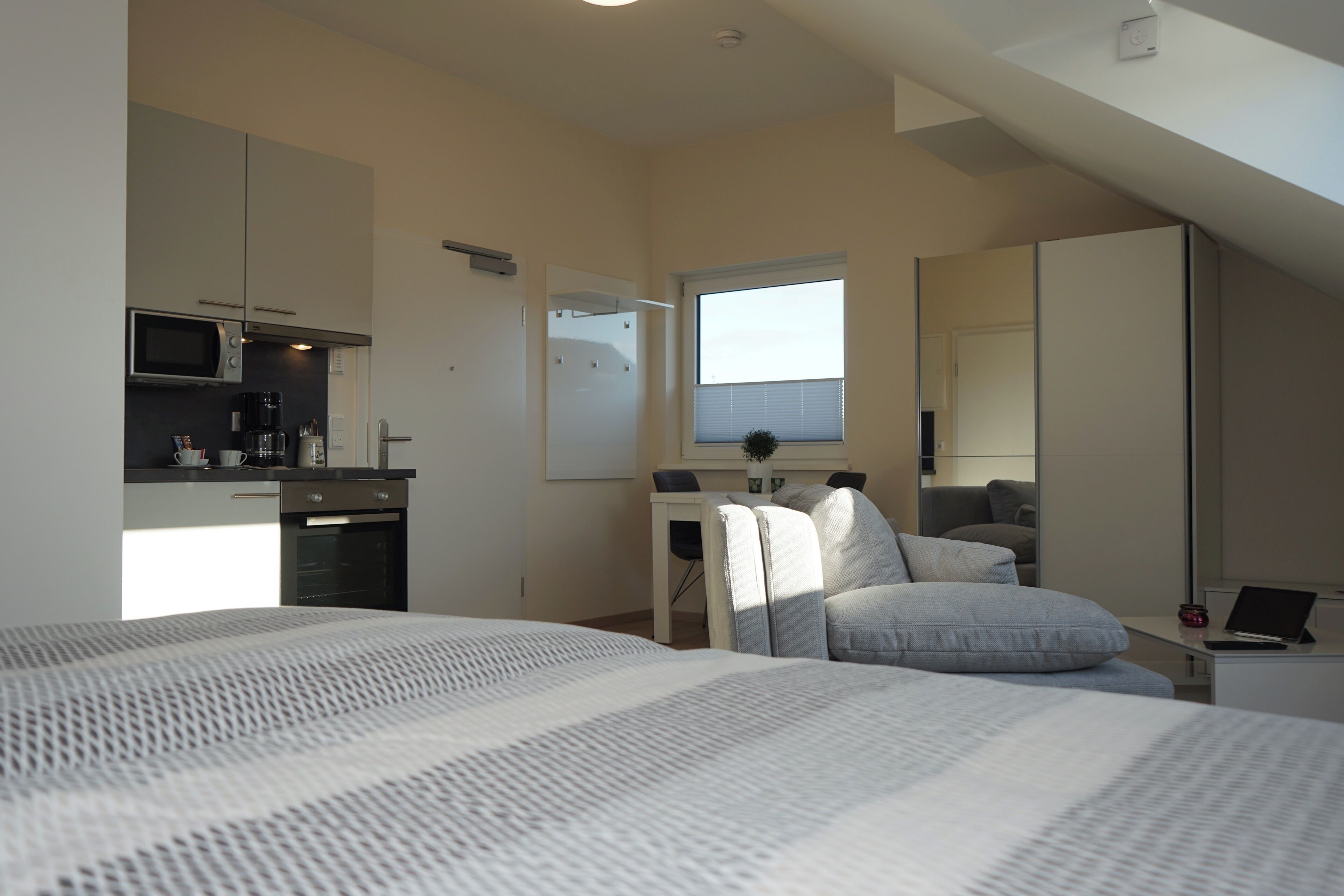 Een compacte slaapkamer met een bed, bureau, stoel, een kleine kitchenette en een raam.
