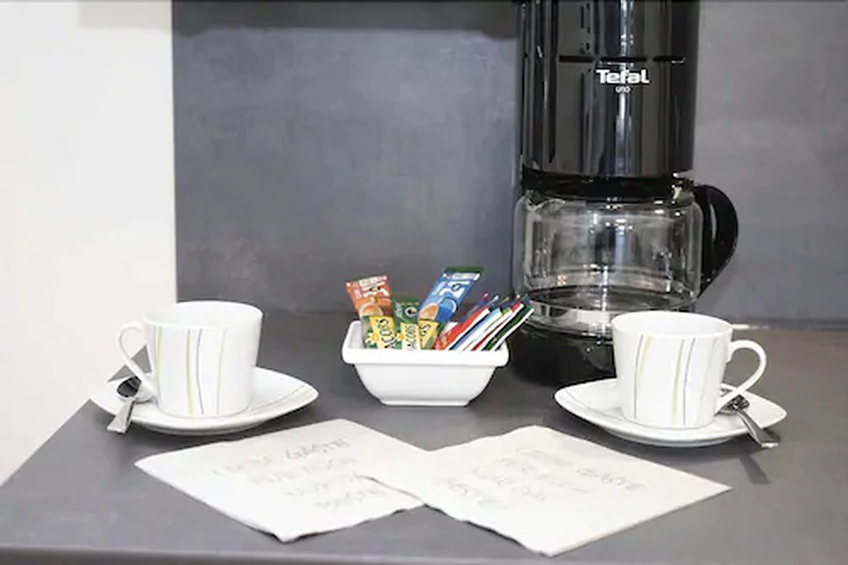 Una estación de café con una cafetera, dos tazas, un bol de cápsulas de café y dos servilletas.