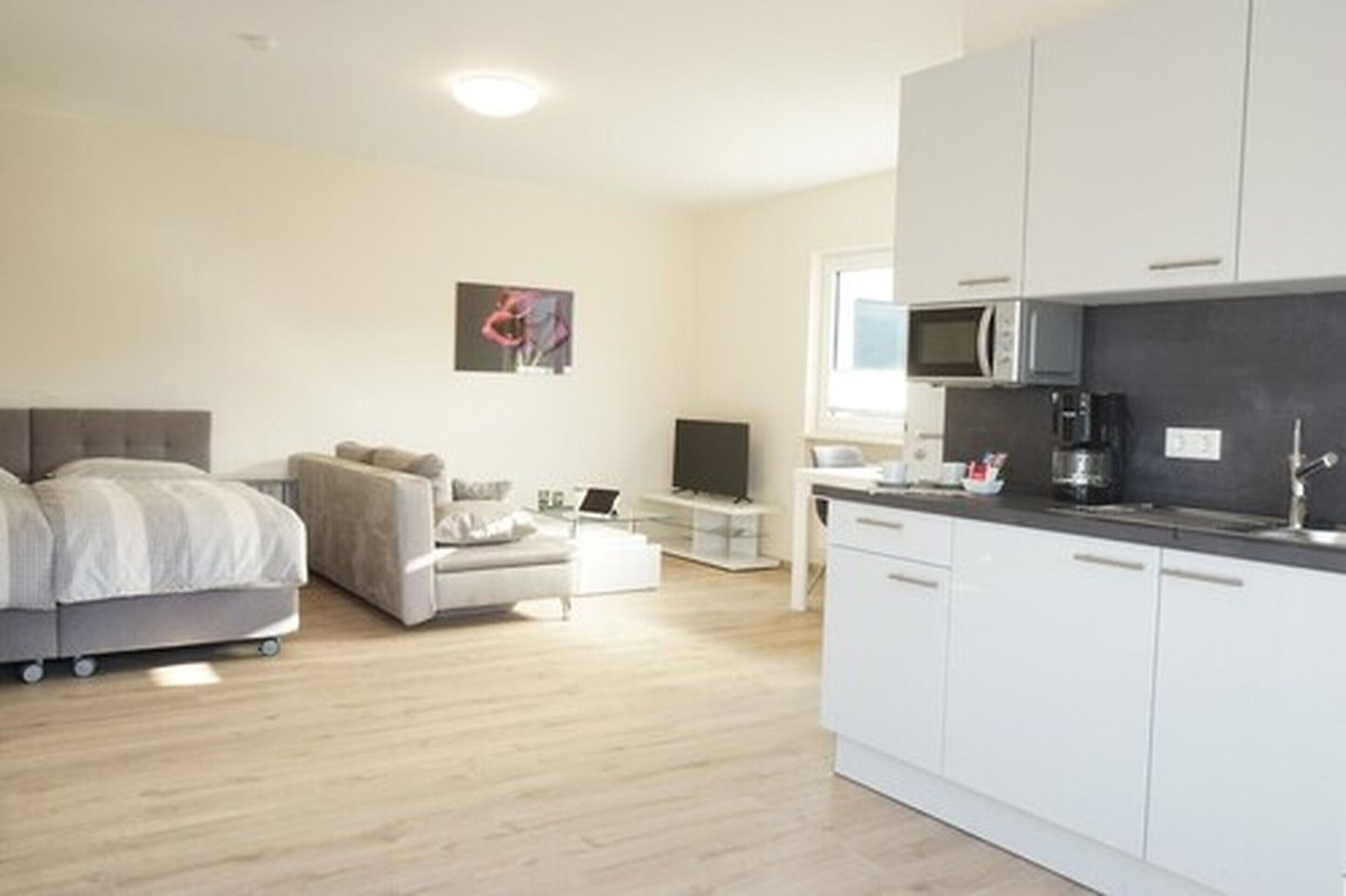 A modern, minimalist living room with a sofa, a small table, and a TV. There is a kitchenette with a microwave and a coffee maker.