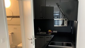 Cocina compacta con microondas, estufa y lavamanos. Una puerta blanca da acceso a un baño con inodoro y cubo de basura.