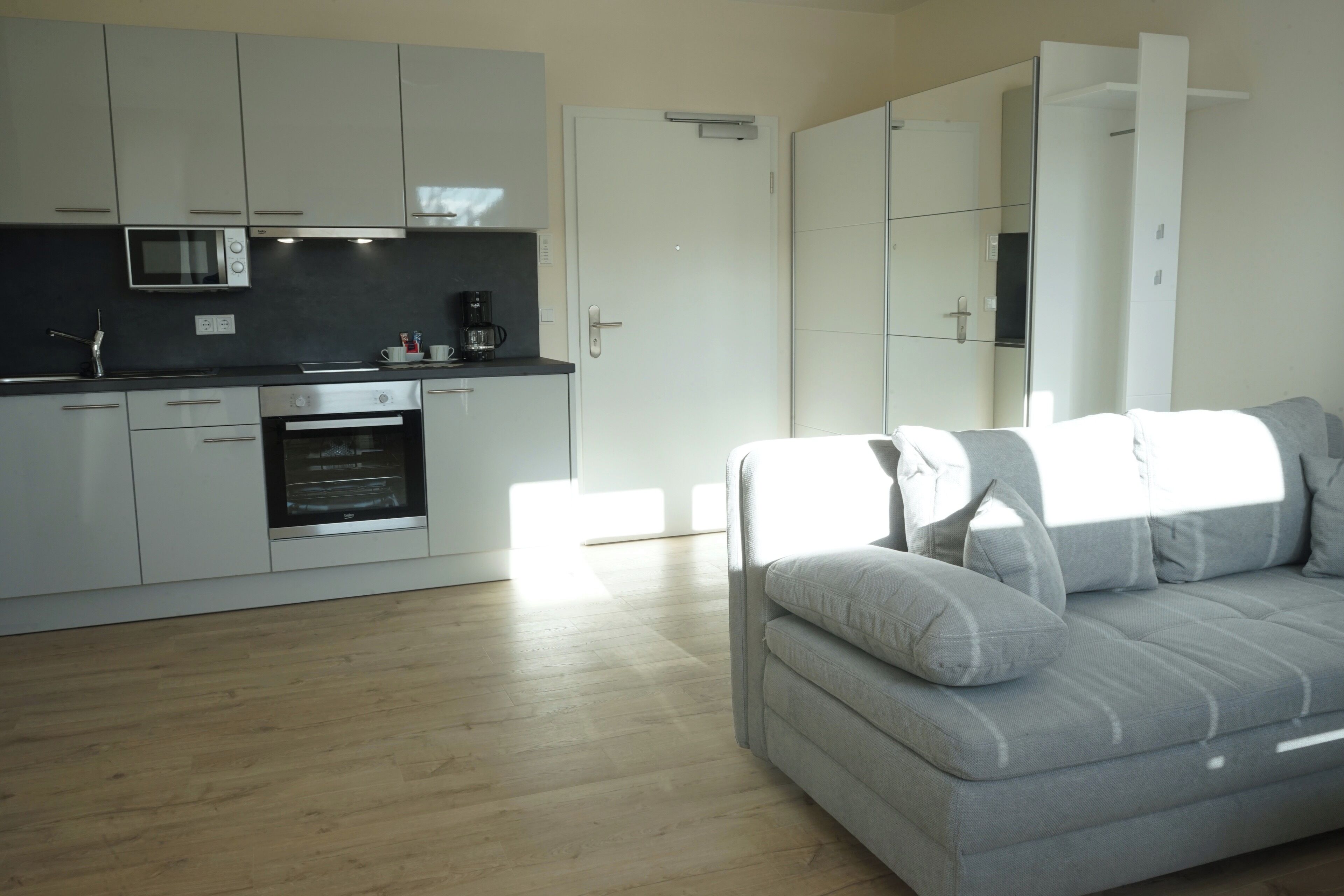 A modern kitchen with built-in appliances and a grey sofa in a bright living area.