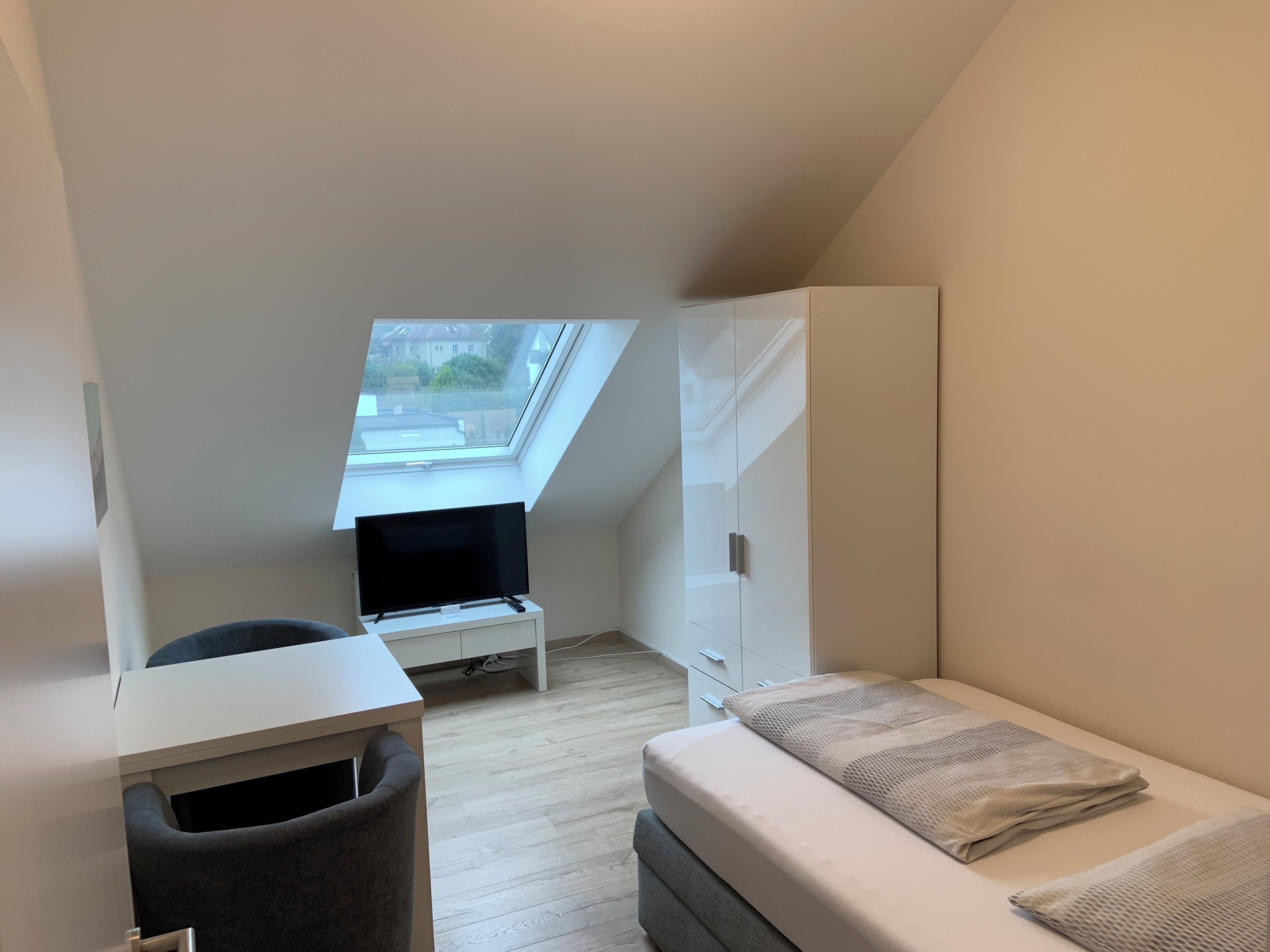 Una habitación pequeña y moderna con techo inclinado, tragaluz, cama, escritorio y armario.