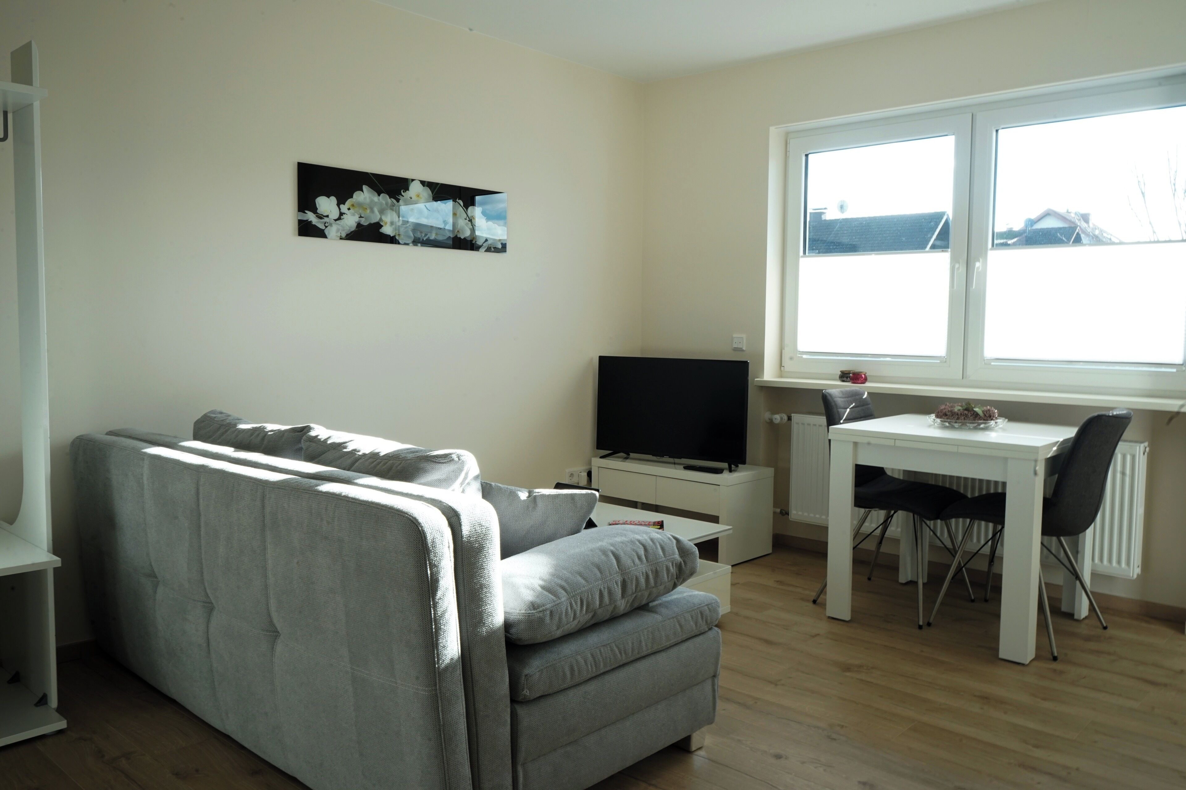 Una sala de estar con un sofá gris, un televisor de pantalla plana, una mesa de comedor con dos sillas y un ventanal con vistas a edificios.