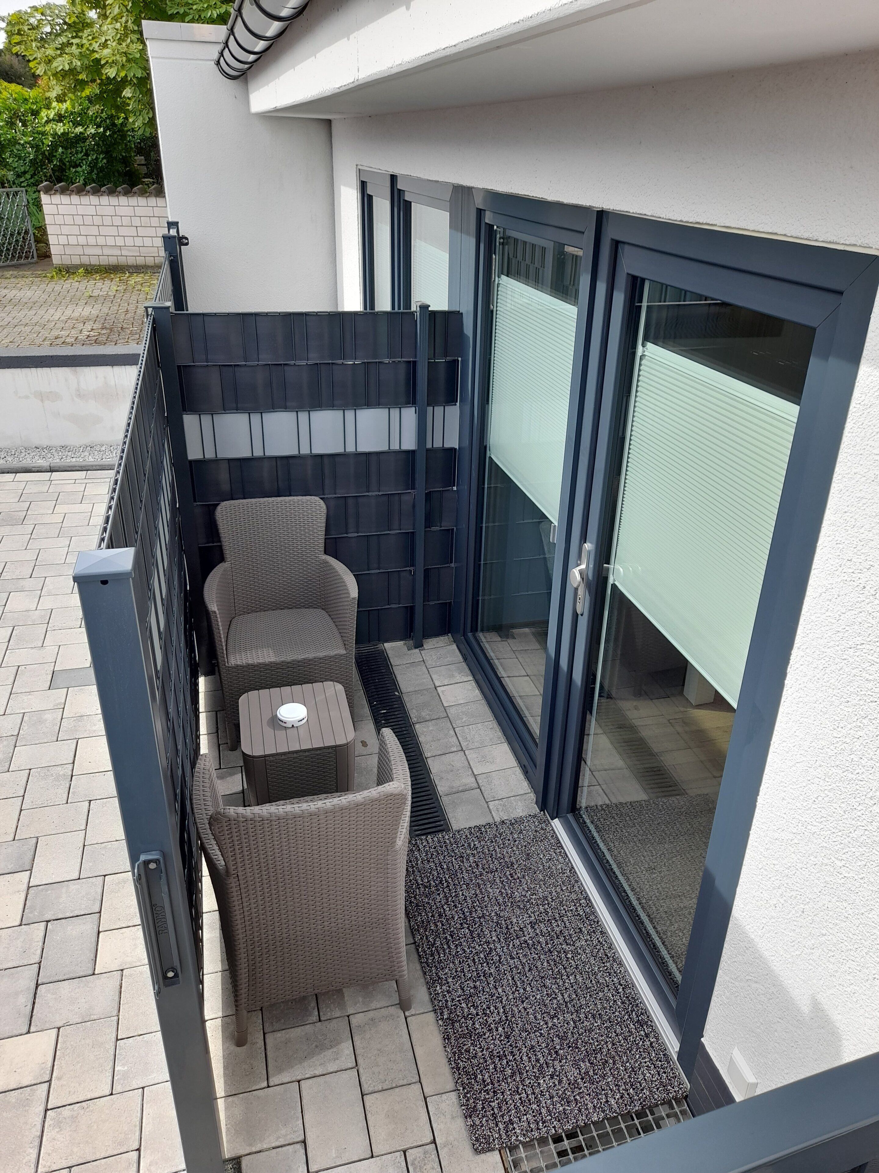 En balkon med fletningsmøbler, et lille bord og en glasdør, der fører til et overdækket område.