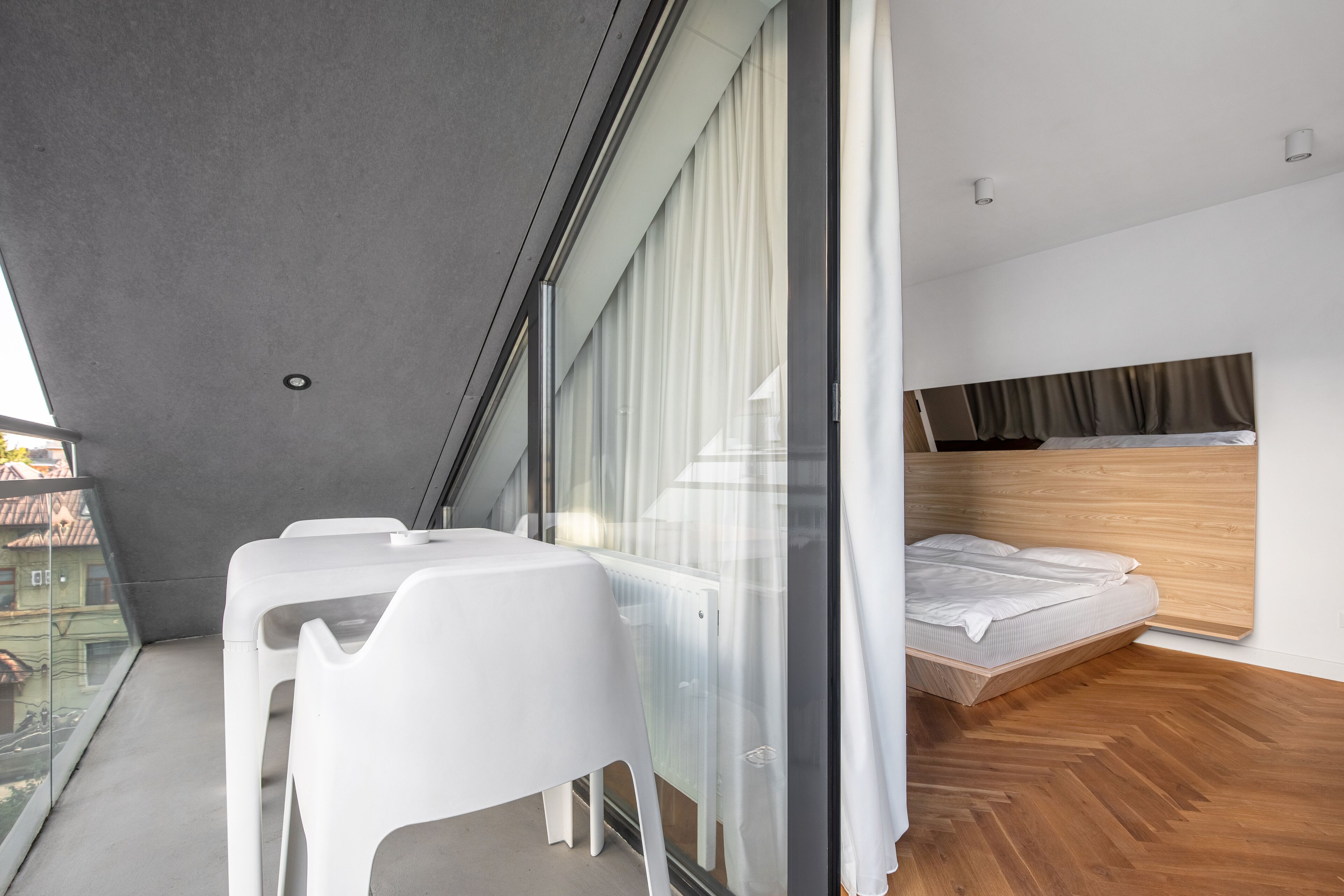 A modern hotel room with a bed, a dining table with chairs, and a view through a large window.