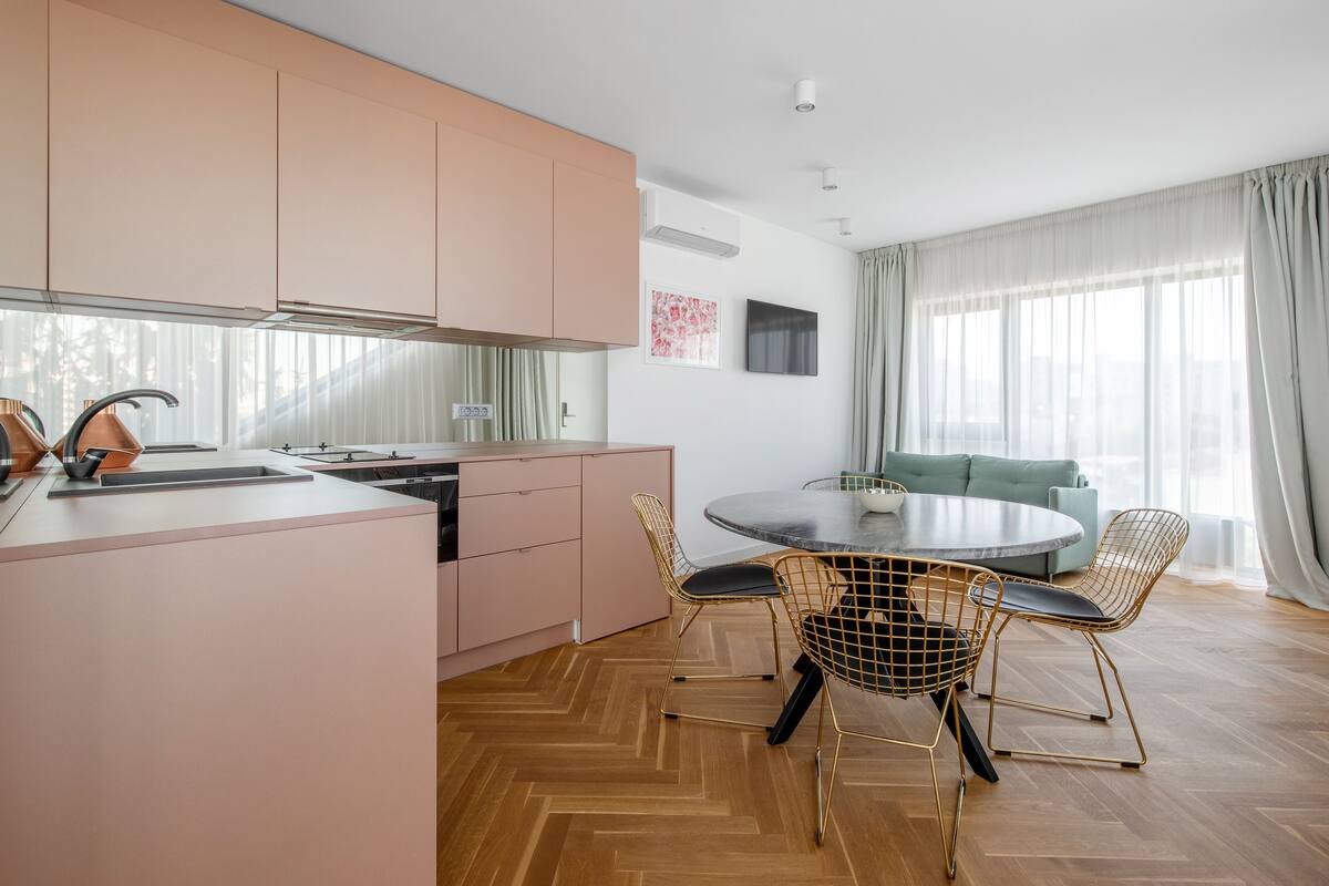 A modern kitchen with a central island, a dining area with a round table and wire chairs, and a living area with a sofa and large windows.