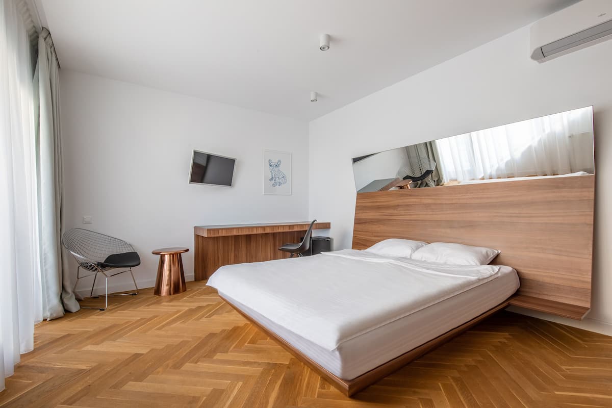 A modern bedroom with a large bed, a wooden headboard, a small desk, a chair, and a television on the wall.