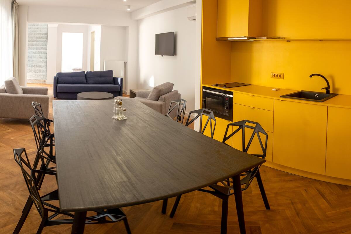 A modern kitchen with a built-in oven and microwave, a dining area with a long table and chairs, and a living area with a sofa and chairs.