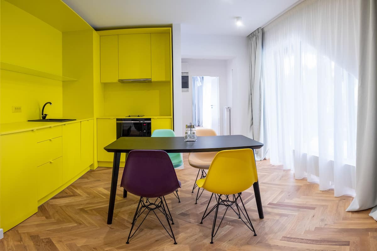 A bright yellow kitchen with a modern stove, a dining table with colorful chairs, and a view of a white room through sheer curtains.