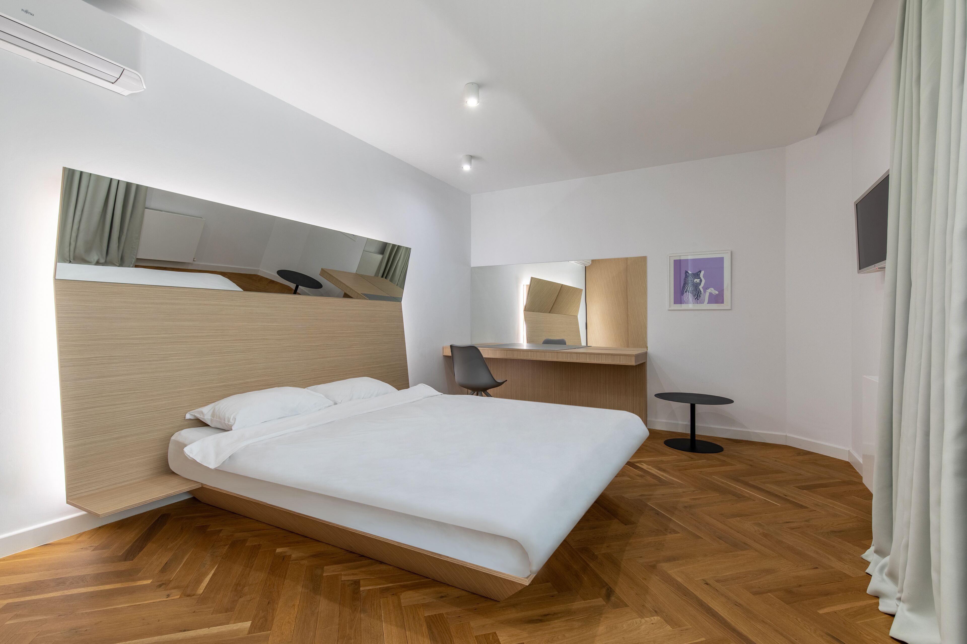 Une chambre d’hôtel moderne avec un plancher en bois, un lit recouvert de draps blancs, un bureau avec une chaise, une petite table ronde et un miroir au mur.