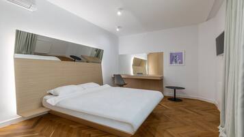 Une chambre d’hôtel moderne avec un plancher en bois, un lit recouvert de draps blancs, un bureau avec une chaise, une petite table ronde et un miroir au mur.