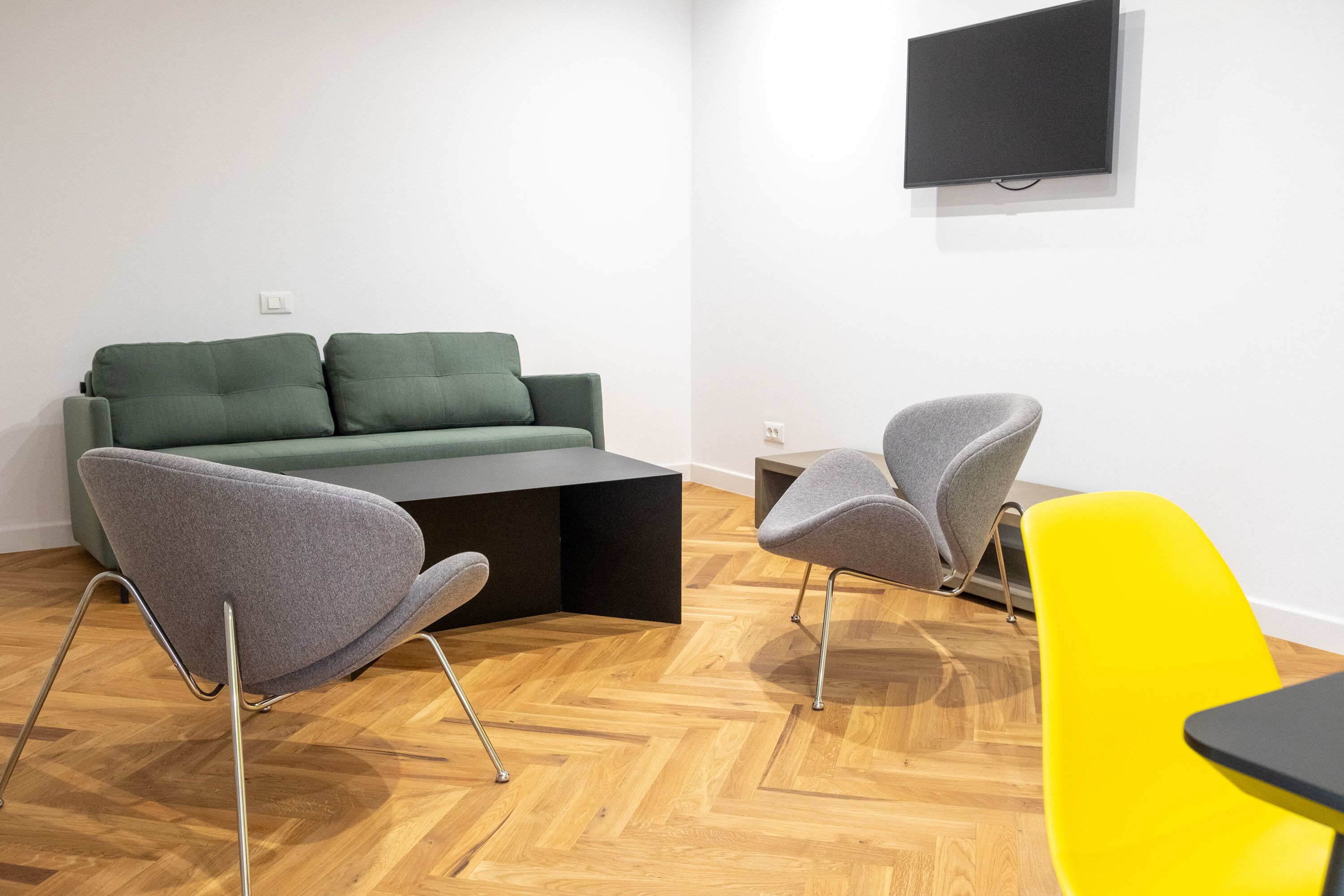 Una sala moderna con un sofá verde, dos sillones grises, una butaca amarilla, una mesa de centro negra y un televisor de pantalla plana montado en la pared.
