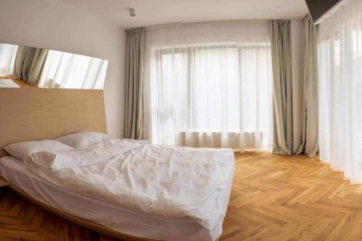 A bedroom with a wooden floor, a bed with white bedding, and sheer curtains.